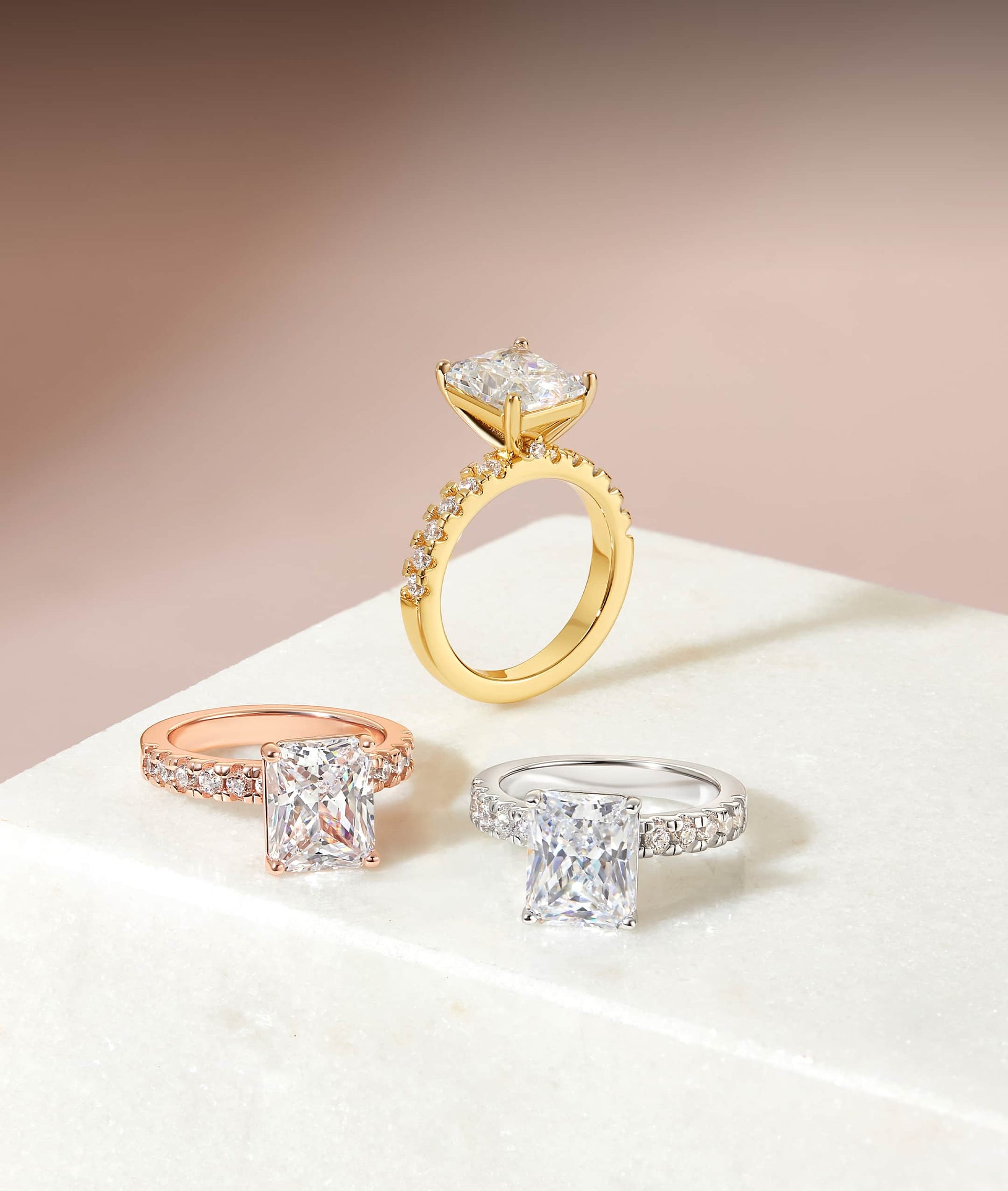 flatlay with gold, rose gold, and silver radiant cut engagement rings with soft pink background