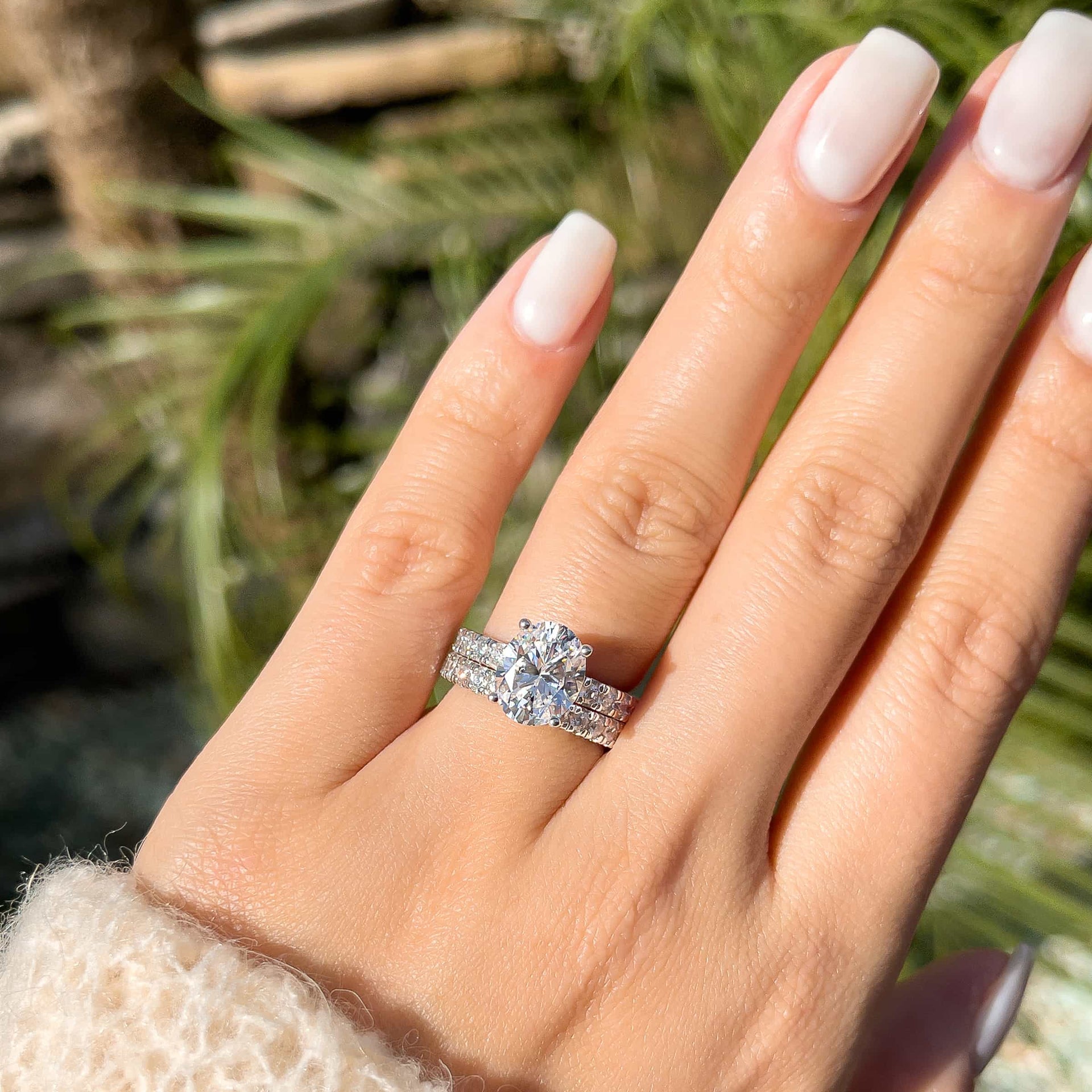 modern silver oval cut engagement ring paired with an eternity band on female hand in front of plants