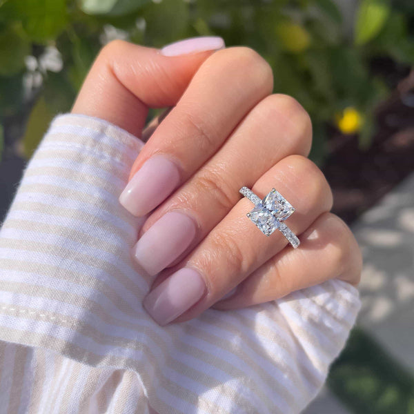 3 carat engagement ring shown in silver on female hand with light pink nails and a striped top in front of some greenery