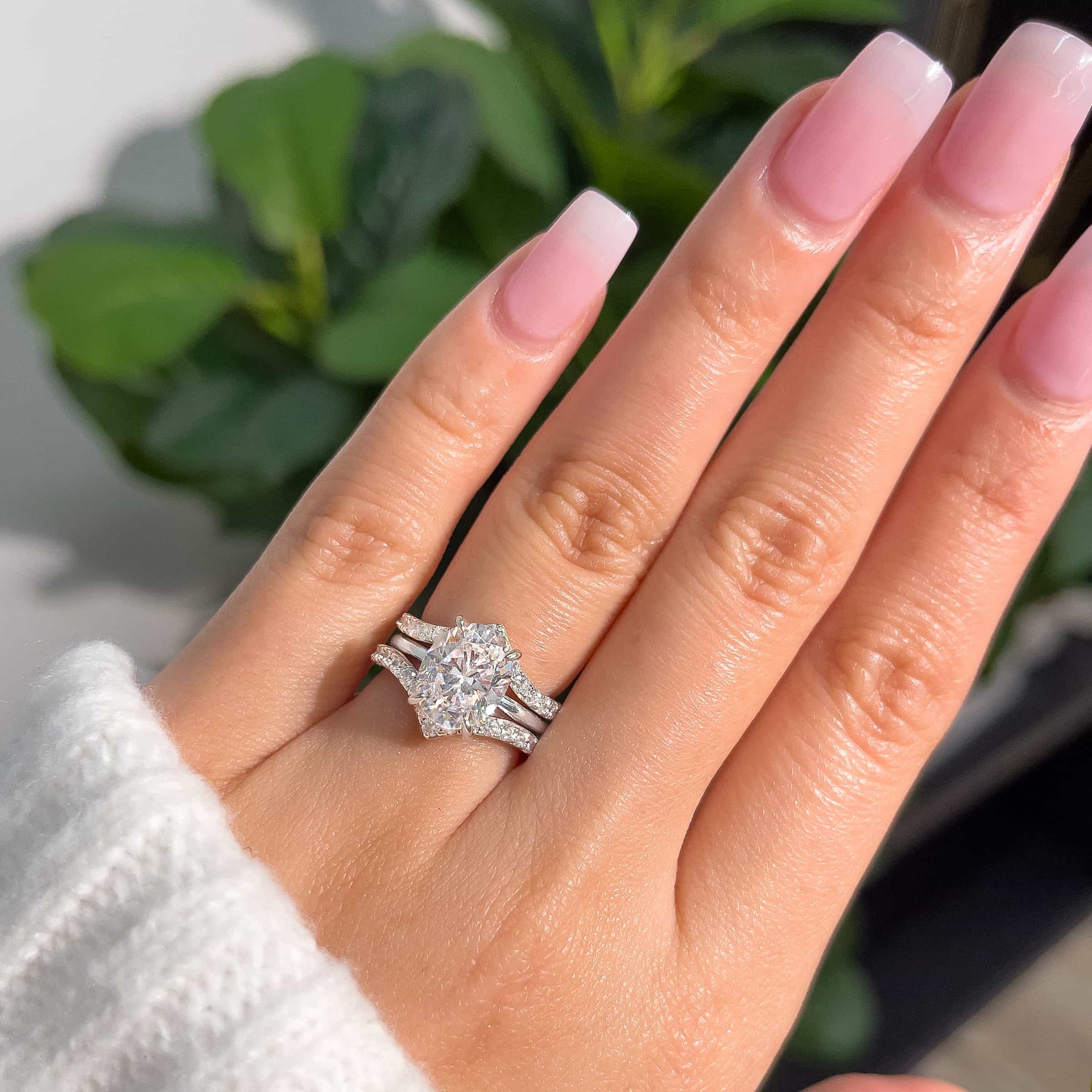 half eternity v-shaped wedding band set of two paired with a 3 carat oval cut solitaire engagement ring modeled on female hand with neutral pink nails