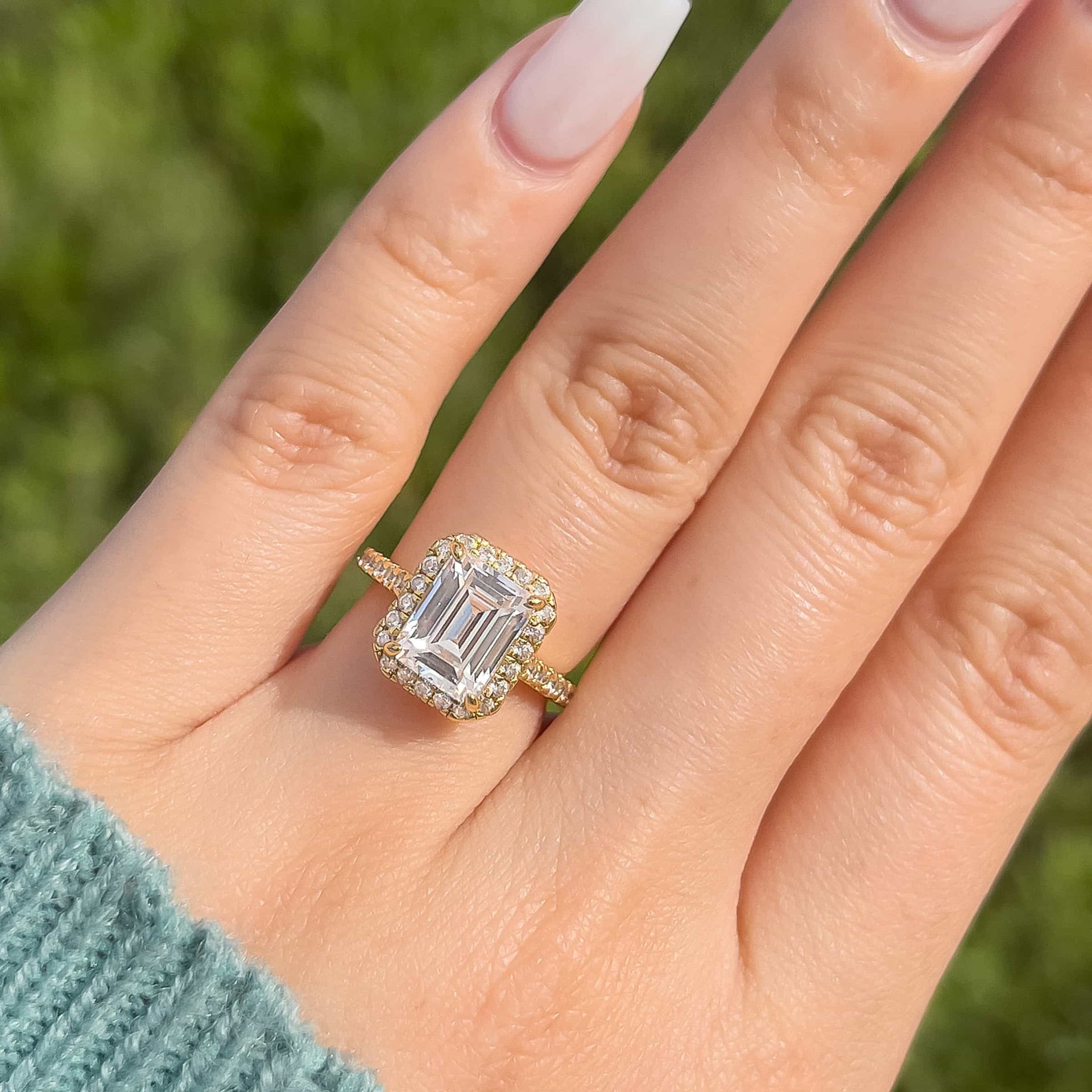 Close up of gold 3 carat emerald cut engagement ring with halo and half eternity band modeled on female hand with teal sweater