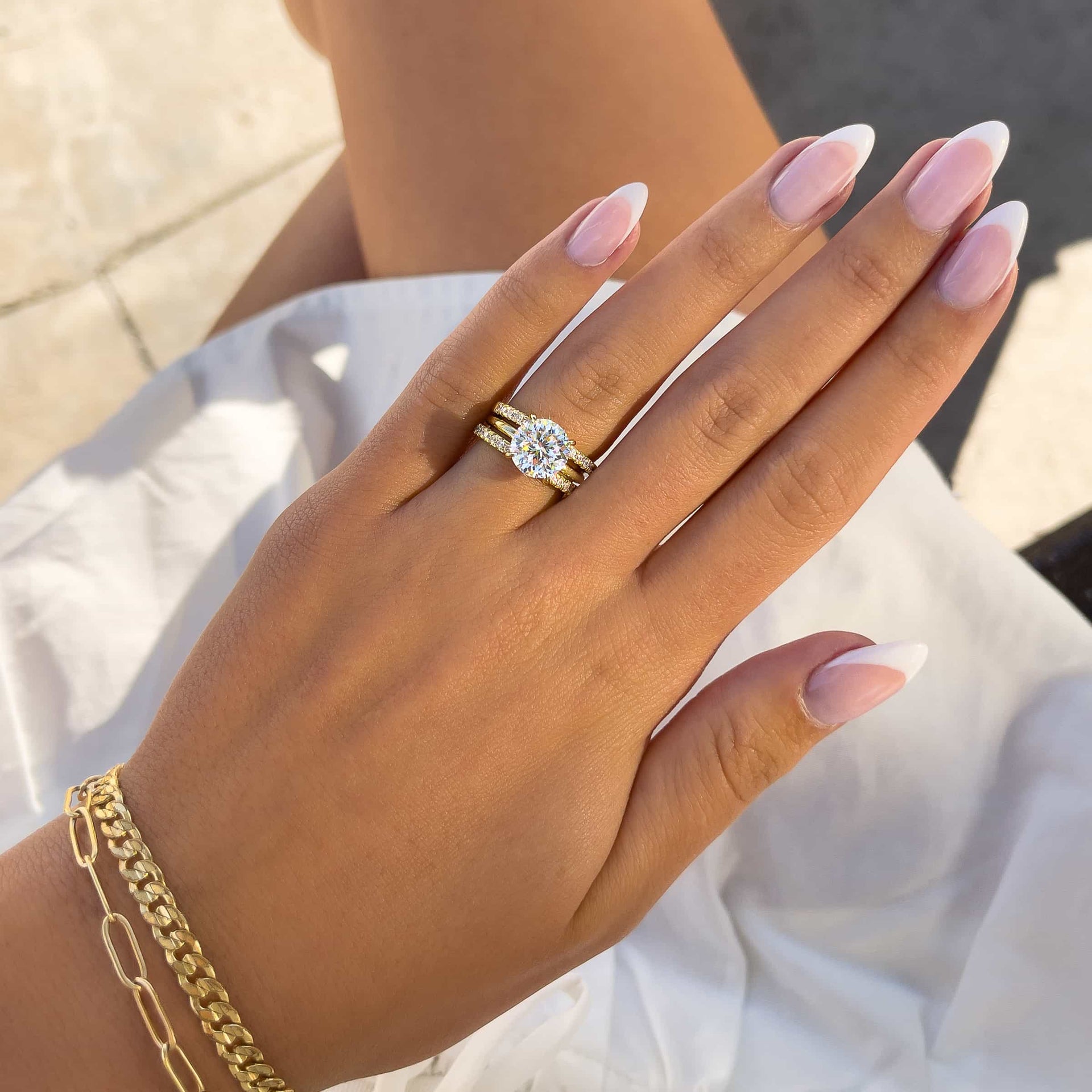 woman wearing round cut engagement ring with two gold wedding bands and gold bracelets with white skirt