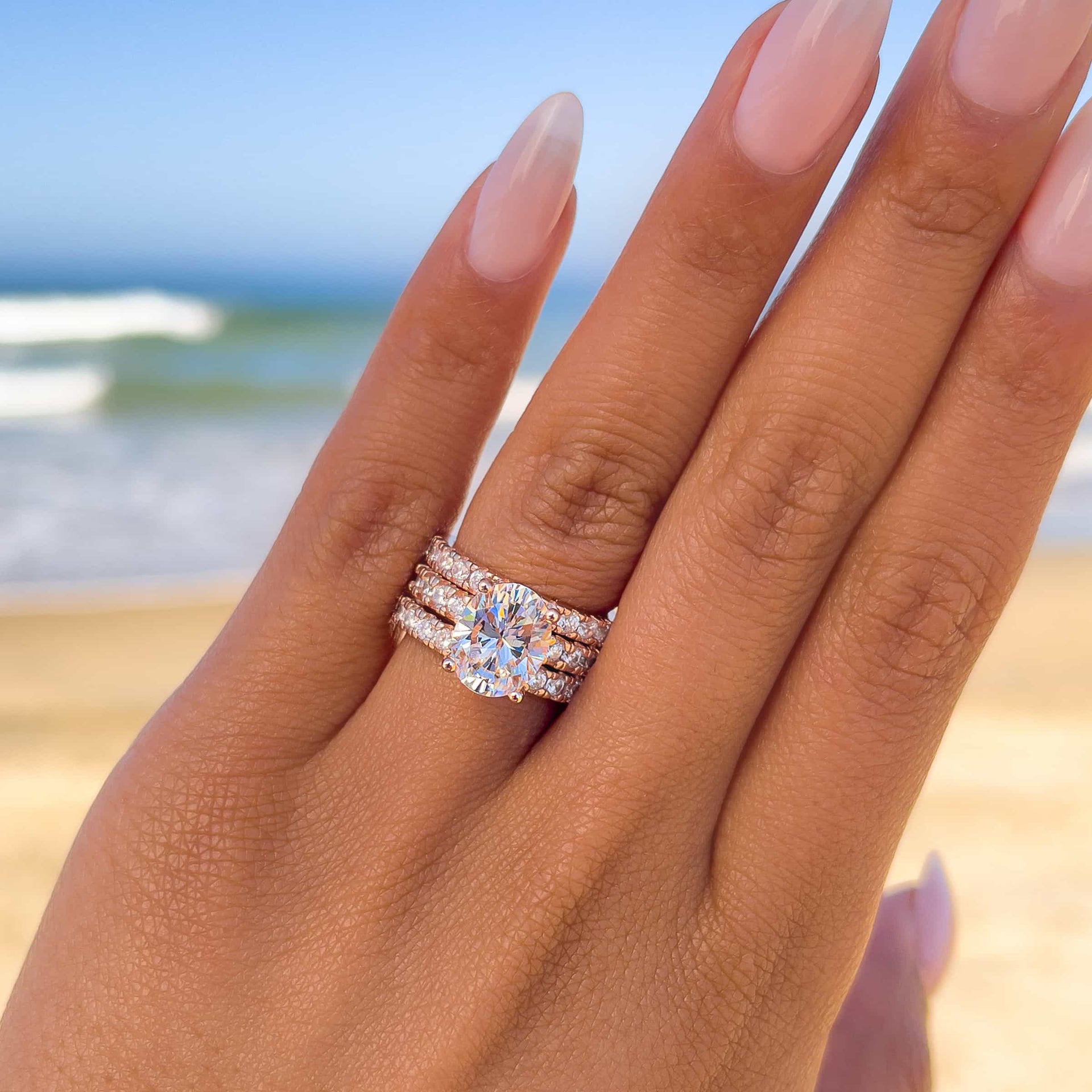 stunning rose gold triple wedding ring stack on model with neutral nails and a coastal background