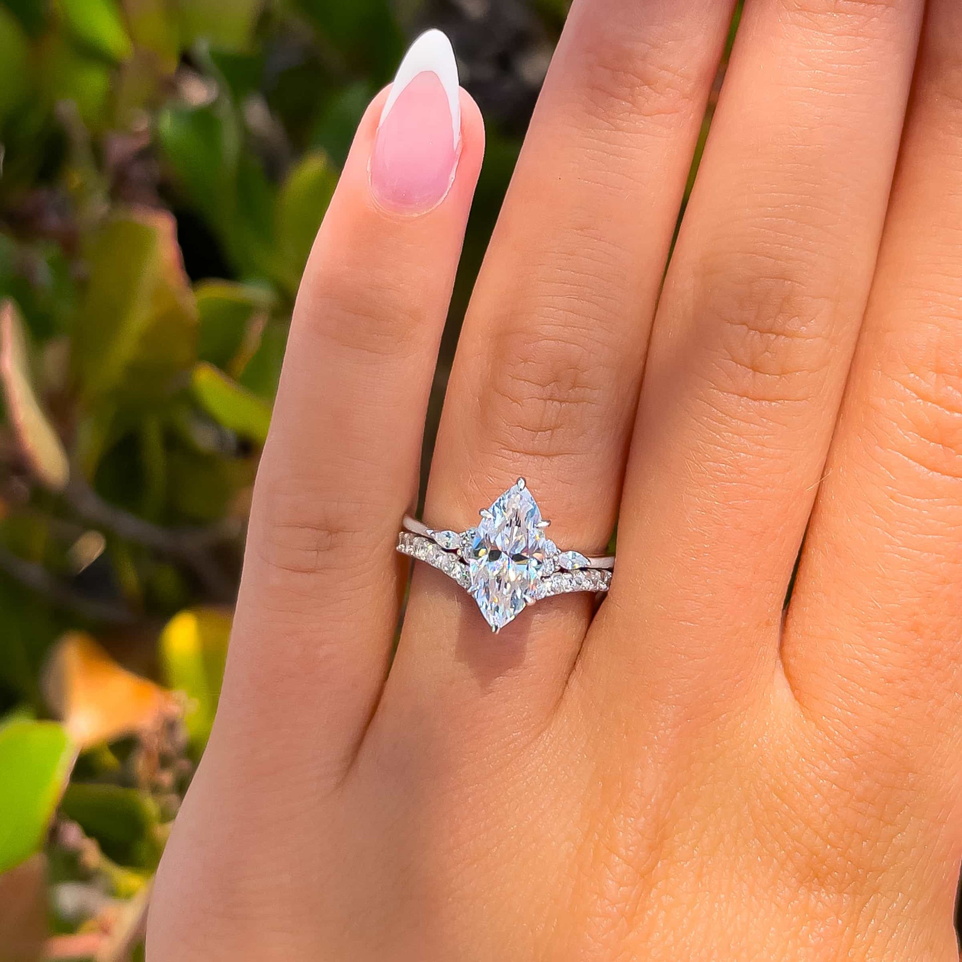 Marquise engagement ring shown on ladies hand with greenery in background stacked with silver wedding band