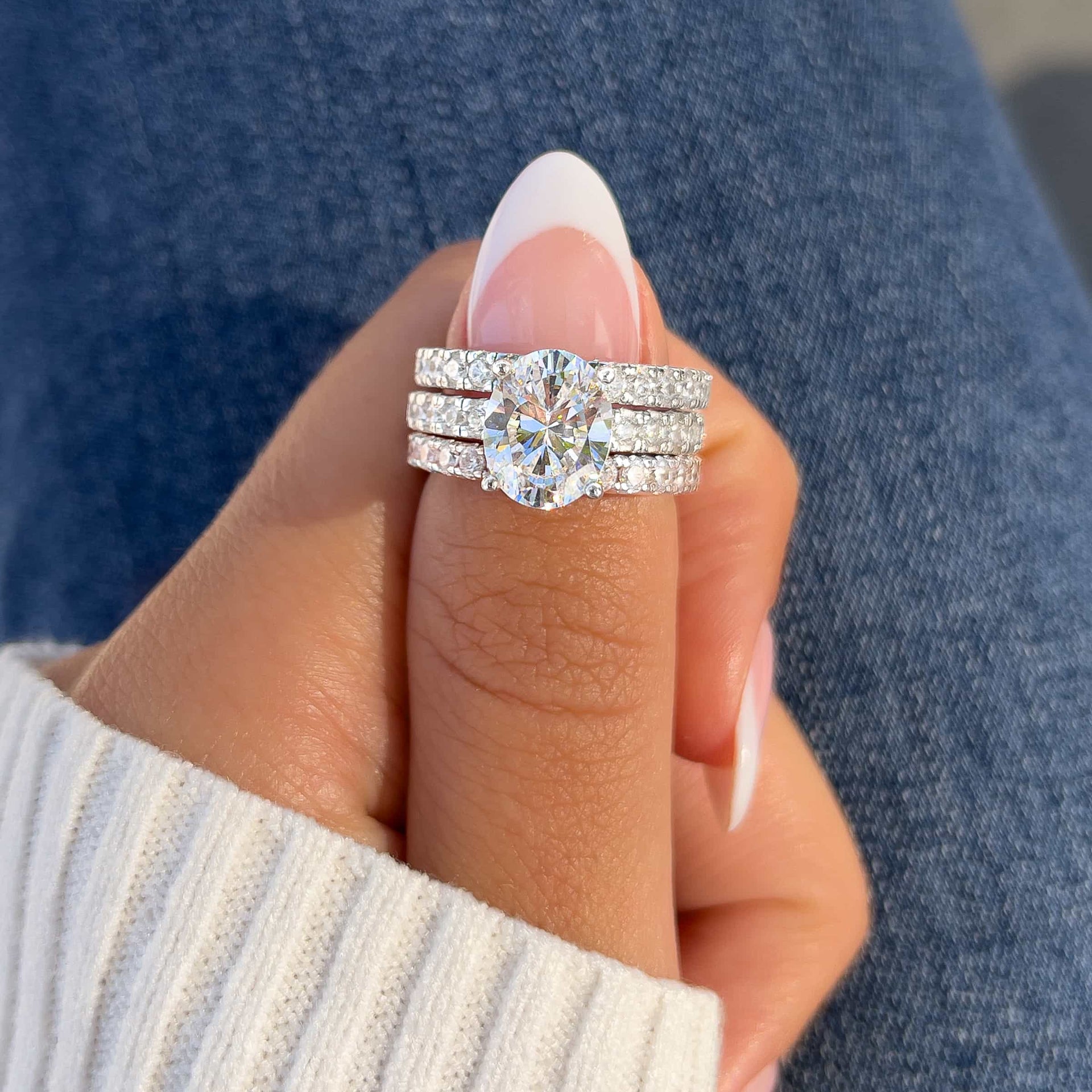 silver triple wedding ring stack made of two silver eternity bands and an oval cut engagement ring on a female model with french tip nails , white sweater, and blue denim