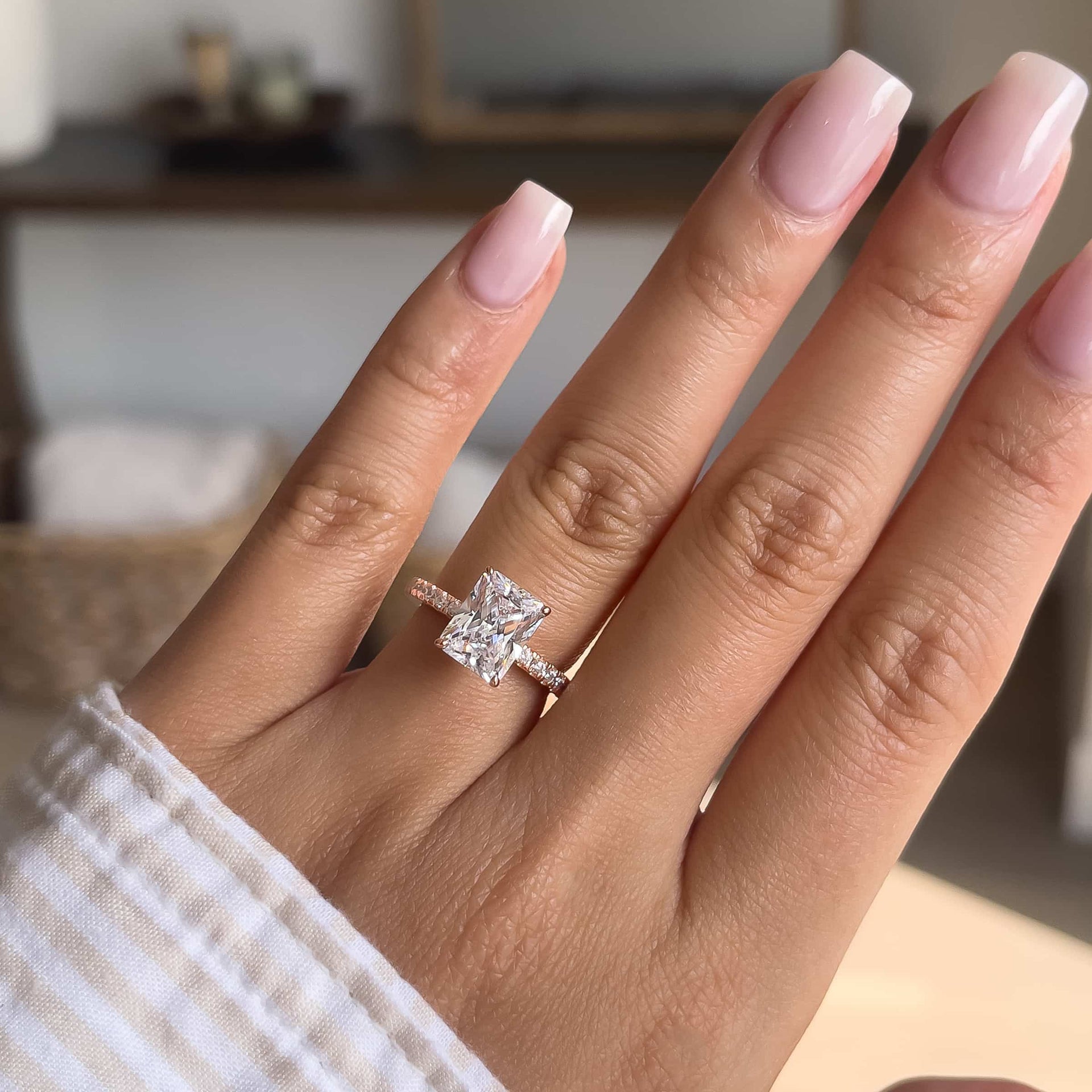 3 carat engagement ring shown in rose gold on female hand with light pink nails and a striped top with a neutral toned background