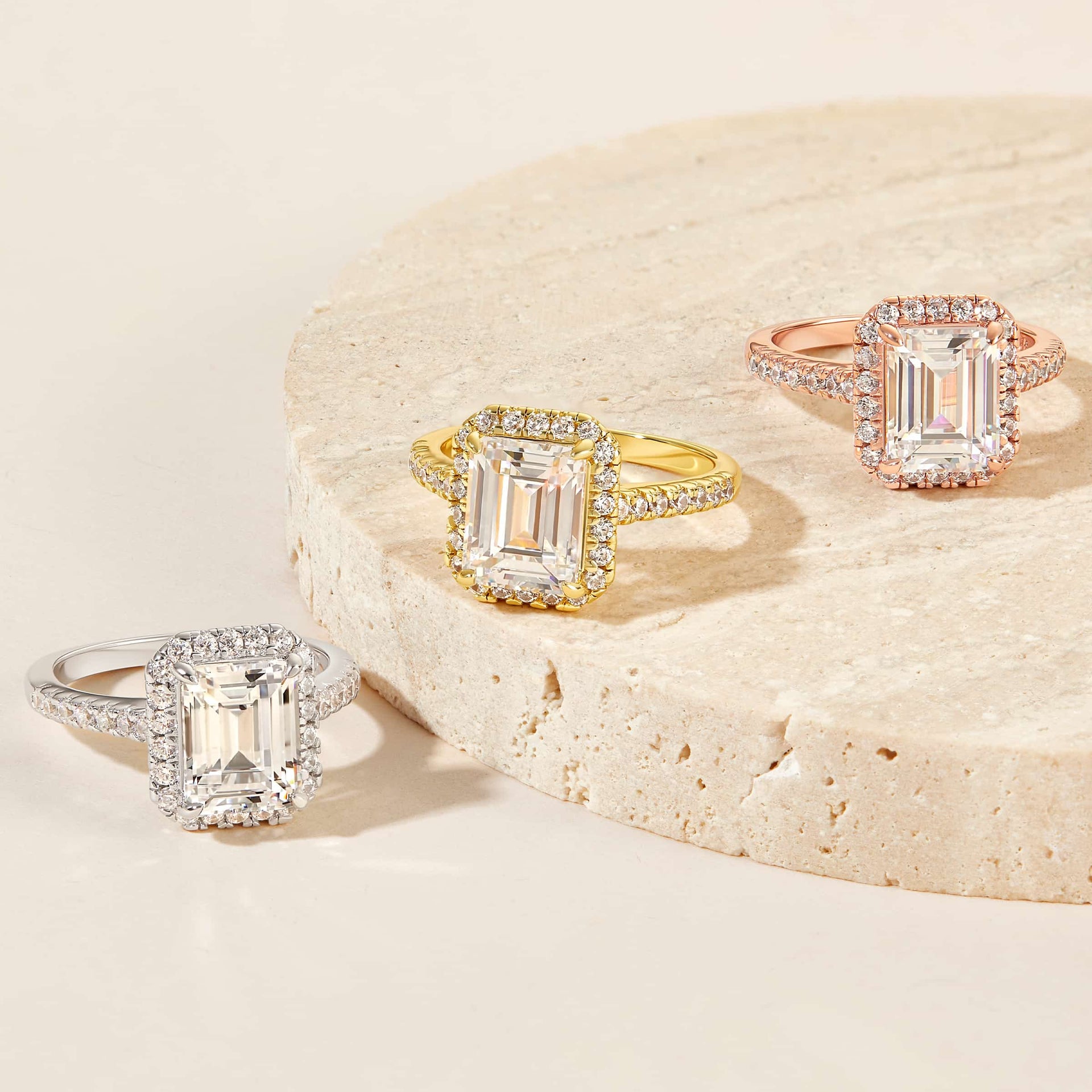3 carat emerald cut engagement ring with halo and sparkly half eternity band shown in its three variants of silver, gold, and rose gold on a neutral stone background