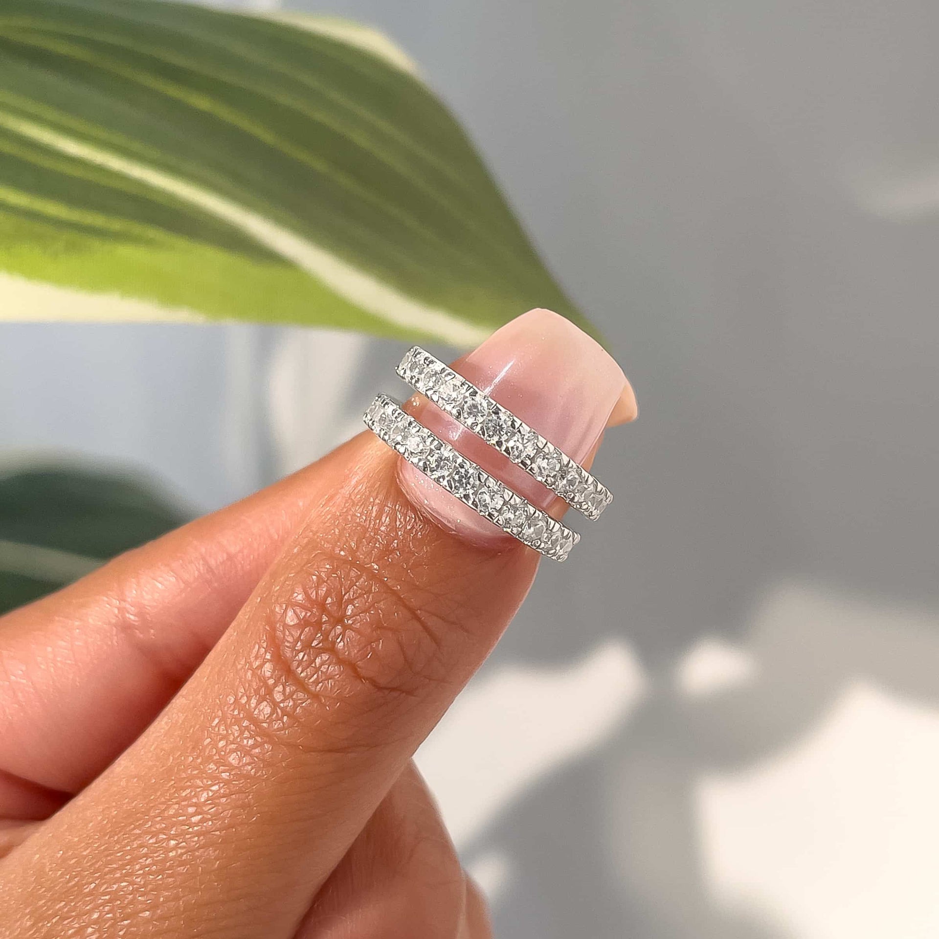 two 2.6mm silver eternity bands being held on a thumb with neutral pink nails in front of a white shadowy wall and greenery
