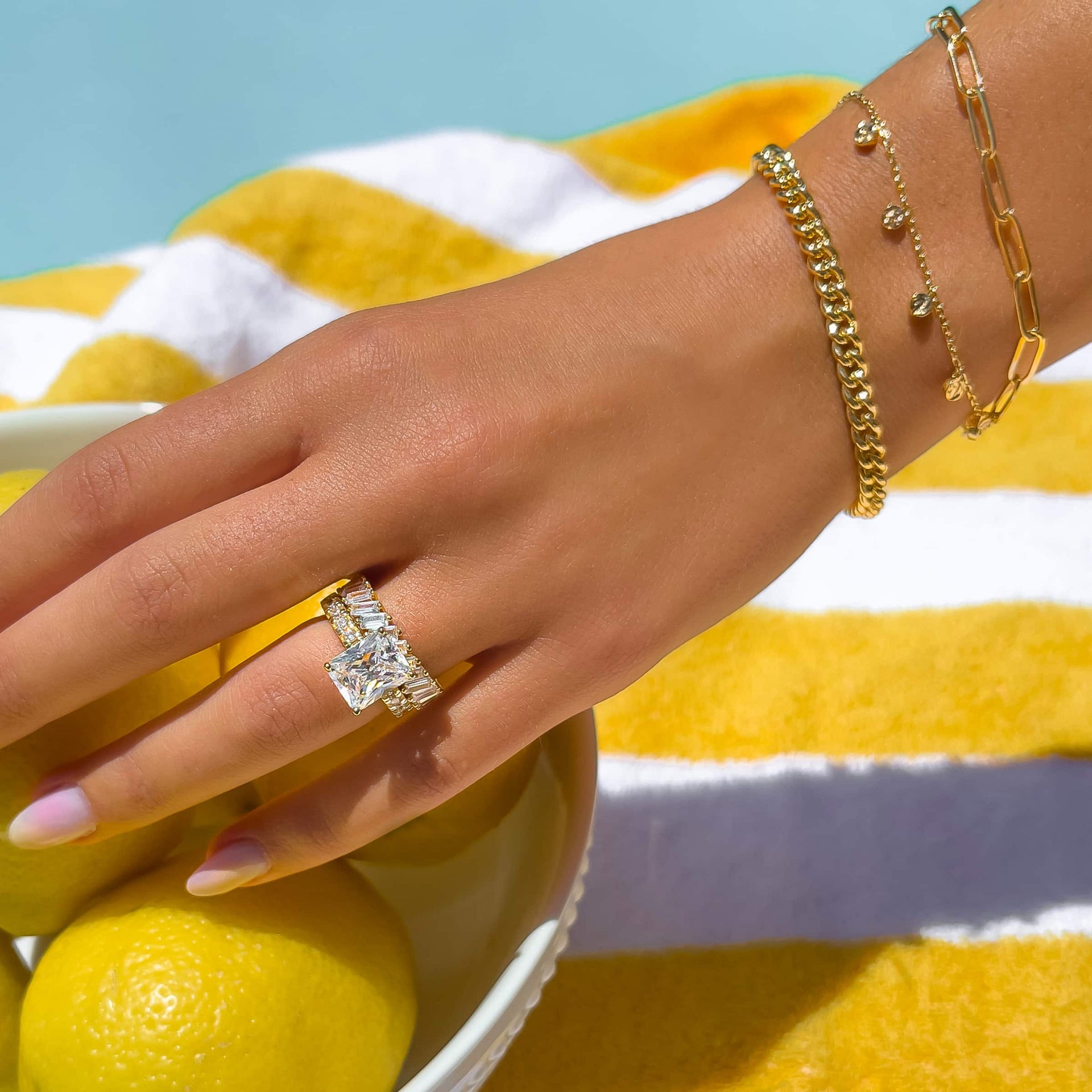 gold radiant cut engagement ring paired with unique baguette engagement ring on model also wearing gold bracelets who is poolside on a striped yellow towel