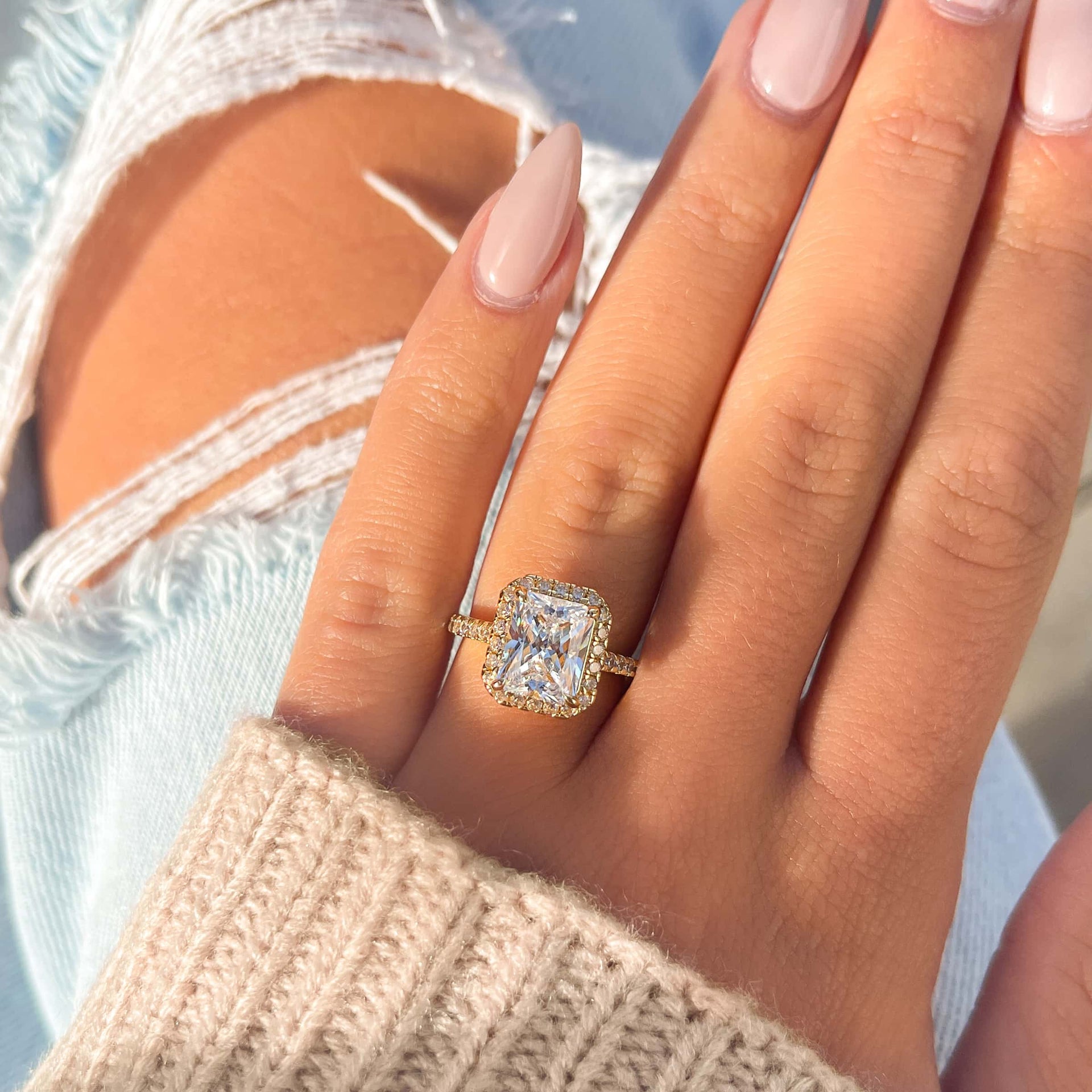 gold halo radiant engagement ring on female hand with neutral sweater and ripped light wash jeans