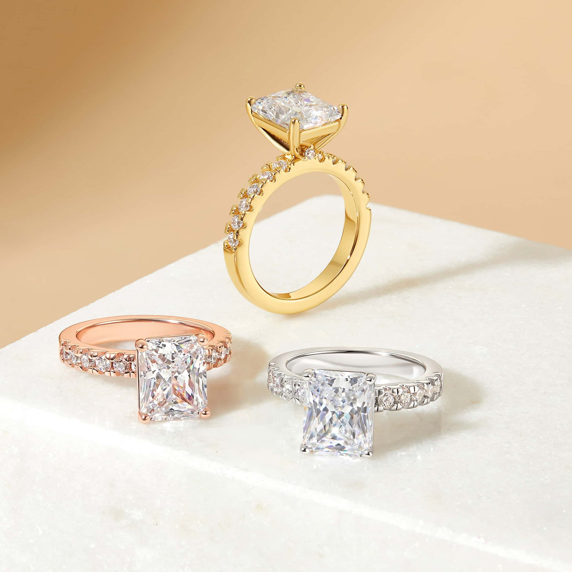 3.75 carat radiant cut engagement ring with sparkly half eternity band shown in its three variants of silver, gold, and rose gold on a neutral stone background
