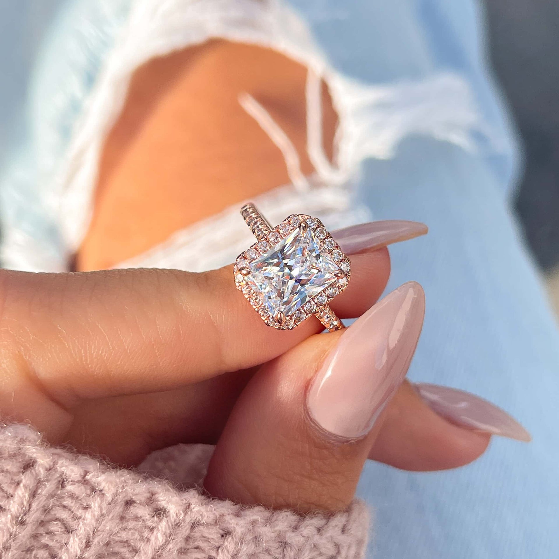 classic rose gold radiant cut halo engagement ring modeled on female hand with pink sweater and ripped light wash jeans