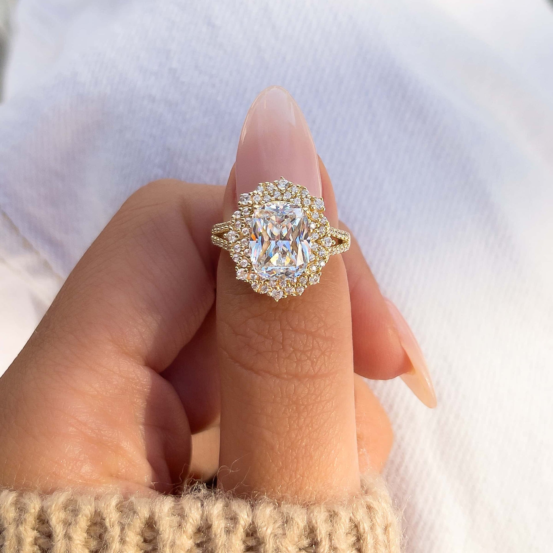 woman pinching gold elongated cushion cut engagement ring shown in gold with light pink nails and white pants