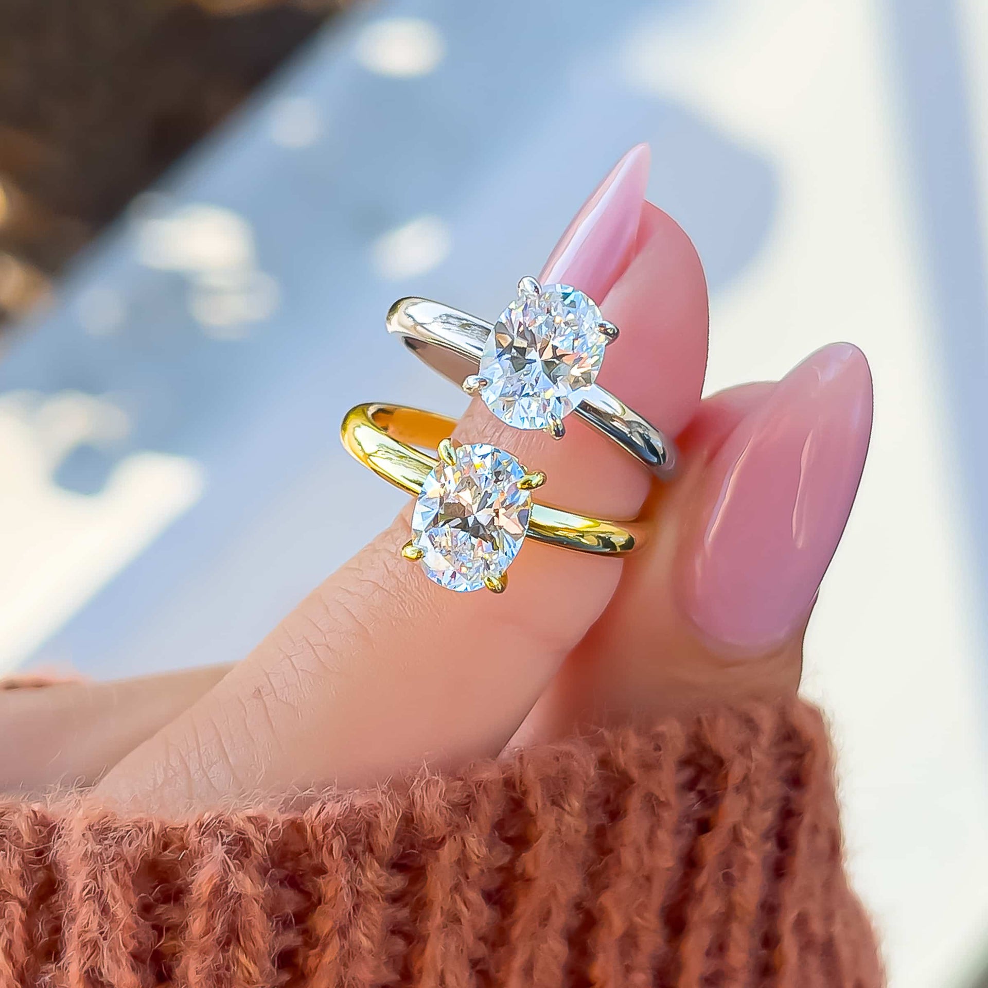 beautiful gold and silver solitaire oval cut engagement rings held by model with pink nails and orange sweater